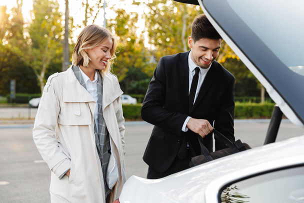 Photo of smiling colleagues man and woman putting bag in car trunk - Photo, Image