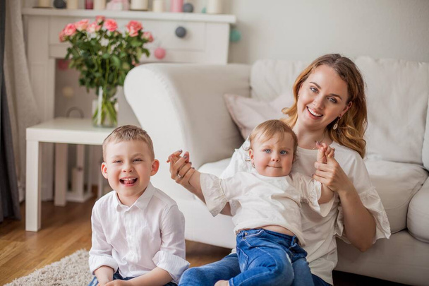 Beautiful young mother, little son, little daughter are sitting on the sofa at home. Mothers Day. A happy family. Cozy. - Fotografie, Obrázek