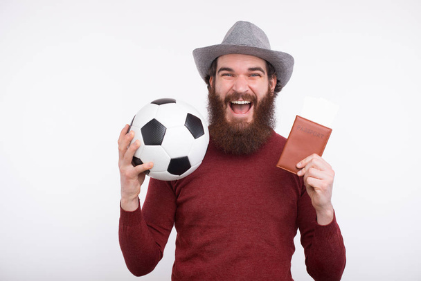 Happy bearded man in casual holding soccer ball and passport with tickets - Photo, Image