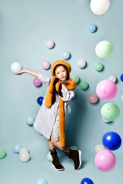 Asian child in oversized shirt dress, brown beret and scarf, boots. She sending air kiss, posing with balloons on blue background. - Foto, afbeelding