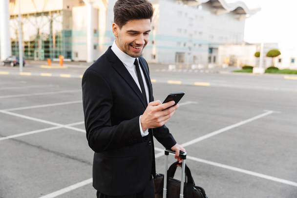 Image of businesslike man standing with luggage and cellphone ou - Foto, afbeelding