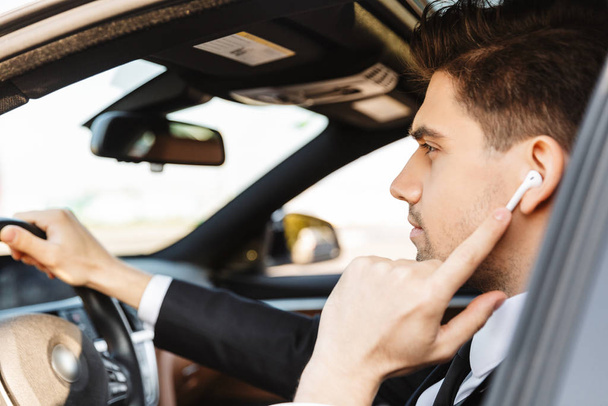 Photo of serious young businessman using earpod while driving car - Photo, Image