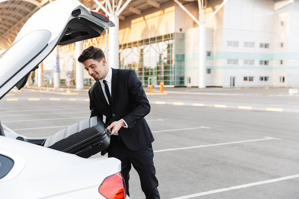 Photo of smiling businessman putting suitcase in car trunk - Foto, imagen