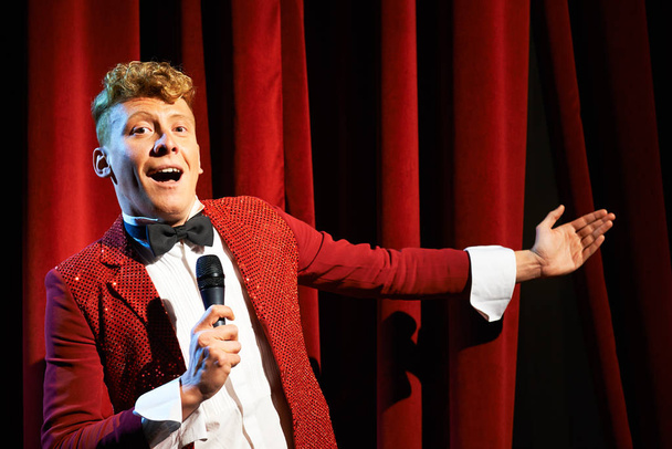 Arts and entertainment in theatre with funny man working as anchorman, standing against red curtains with microphone - Zdjęcie, obraz
