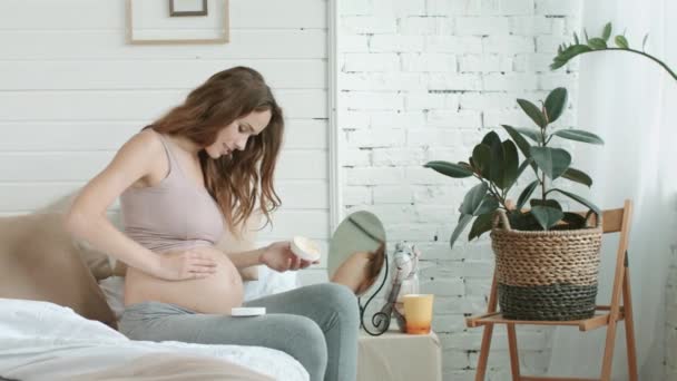 Young pregnant woman applying skincare cream on pregnant belly in bedroom. - Πλάνα, βίντεο
