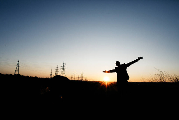 Mann breitet seine Hände auf Stromleitungen aus sparen Sie den Planeten und die Elektrizität. Freiheit fühlen. Stromverteilungsnetz - Strommasten gegen orangen und gelben Sonnenuntergang. die Eiffeltürme - Foto, Bild