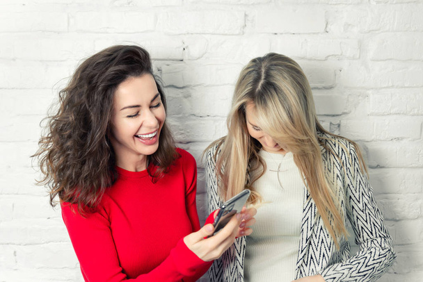 Two young adult caucasian girlfriends having fun and enjoy spending time together against white brick wall on background. Pair of friends smiling making selfie picture for social networks at home - Photo, Image