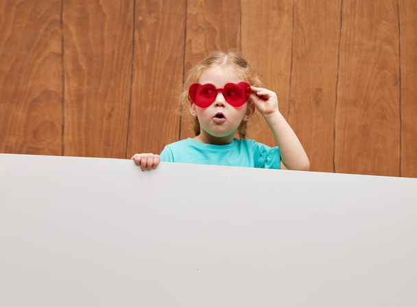 Surprised Little Child girl in heart shap sunglasses peeks out from behind a white banner. Banner. Happy childhood. Wow funny face  - Photo, Image