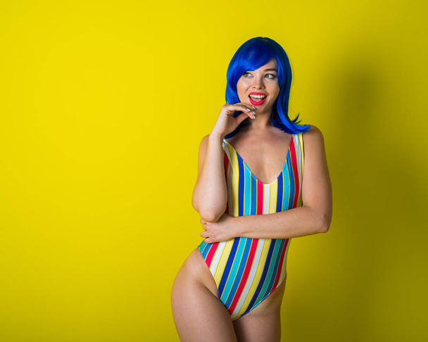 Beautiful woman in a striped swimsuit in a blue wig posing on a yellow background. Portrait of a glamorous girl. - Zdjęcie, obraz