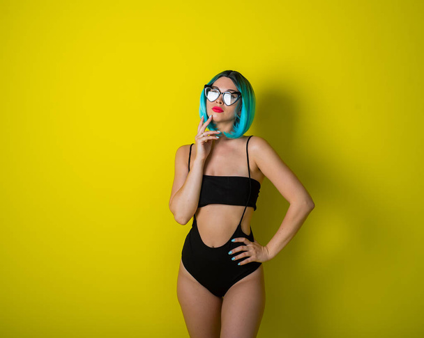 Beautiful woman in a black bikini in a colored wig and sunglasses on a yellow background. Girl posing in the studio. - Foto, imagen