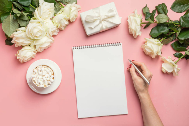 Female hands writing in notebook and bouquet white roses on pink. Top view with copy space. - Foto, Imagem