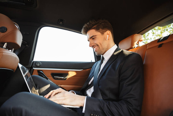 Photo of smiling businessman using earpod and laptop while going in car - Foto, Imagem