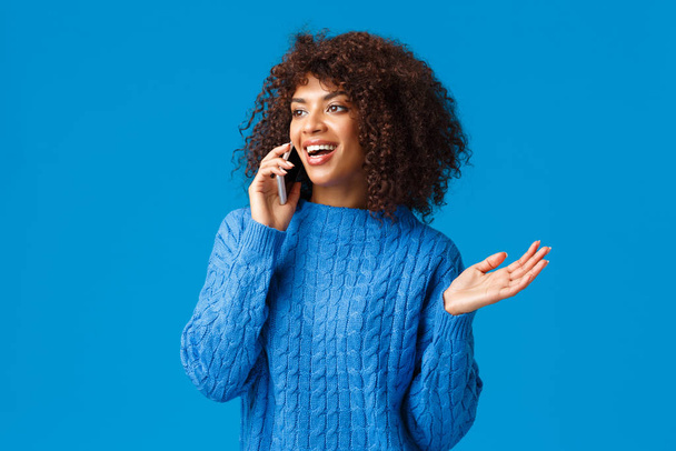 Despreocupado falante atraente mulher afro-americana em camisola de inverno discutir feriados recentes e sua viagem ao exterior ski-resort, falando ao telefone, segurando smartphone perto de gesto ouvido, sorrindo
 - Foto, Imagem