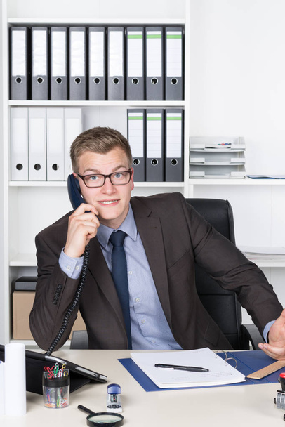 young businessman with glasses and a surprised,shocked expression sitting at desk in the office and on the phone. in the background there is a shelf. the man looks to camera and smiling. - Фото, изображение