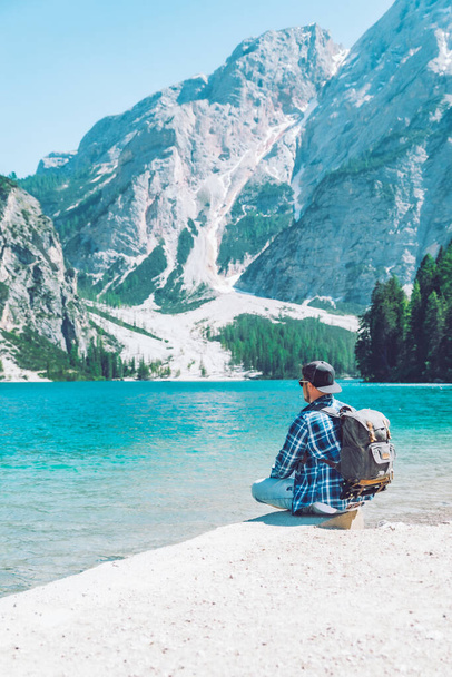 человек сидит на пляже озера с рюкзаком горы на заднем плане
 - Фото, изображение