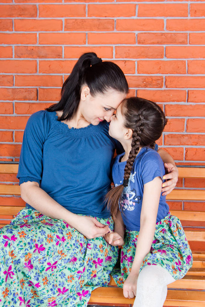 Madre e hija con ropa similar se abrazan y se sientan en el banco frente a la pared de ladrillo rojo. Fotografía de interior
 - Foto, Imagen
