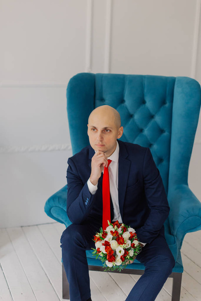 Handsome groom sits on a chair with a wedding bouquet. - Фото, изображение