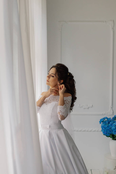 Beautiful bride puts on earrings by the window. Happy wedding day. - Foto, afbeelding