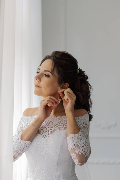 Beautiful bride puts on earrings by the window. Happy wedding day. - Photo, Image