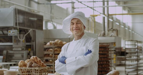 パン屋のキッチンの作業台の横に立っている間に大きな笑みを浮かべてスタイリッシュな制服姿の男のコマーシャル顔のパン屋のカメラの肖像画の前で - 写真・画像