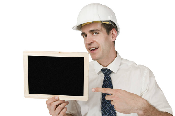frontal torso shot of a young man in white shirt and white helmet a small chalkboard upholding it,pointing against white background happy kchelnd with forefinger with tilted head in the camera  - Photo, image