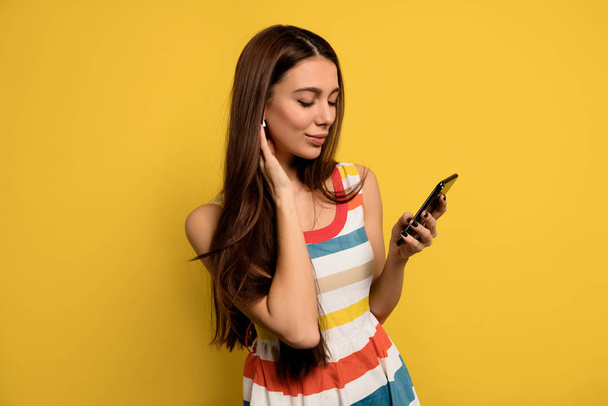 Inside portrait of lovable woman with long hair wearing bright dress holding smartphone and listening music in headphones  - Foto, afbeelding