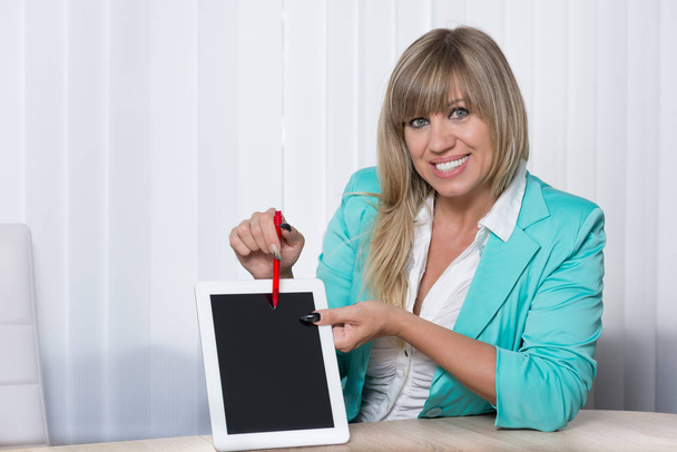 a woman sitting at table in office and pointing with a pen on a tablet. the woman looking to the camera. - Foto, immagini