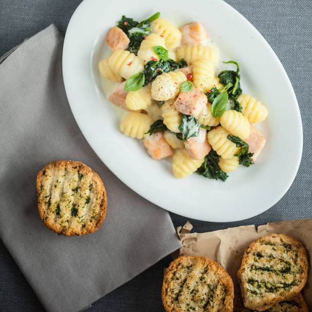 View from above of a plate of Italian gnocchi pasta , basil and grilled salmon pieces served with crisp crusty toasted herbal buns - Photo, Image