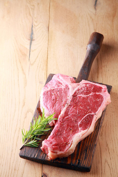 An uncooked tender lean porterhouse steak with a sprig of fresh aromatic rosemary for seasoning on an old rustic wooden board on a wood table with copyspace, view from above - Foto, afbeelding