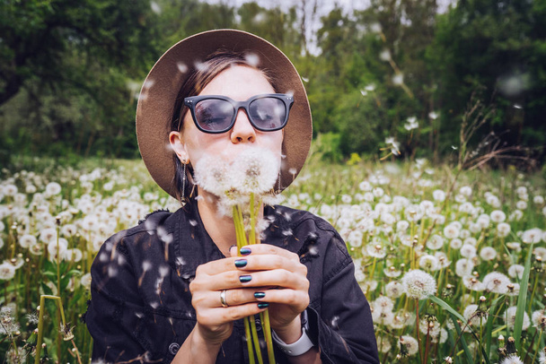 Happy woman blowing on dandelion in park. Girl in hat and sunglasses. Wishing, joy concept - Φωτογραφία, εικόνα