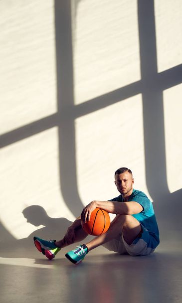 Joven atleta, jugador de baloncesto sosteniendo la pelota con una mano y sentado cerca de la pared con sombras desde la ventana
 - Foto, Imagen