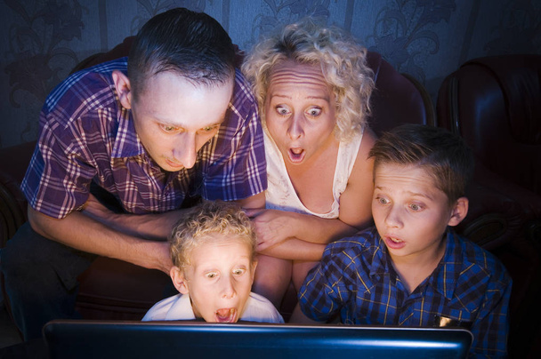 parents with their two sons,ages 7 and 13,sitting in the darkened room on the couch,looking shocked into a monitor towards the camera - Foto, Imagen