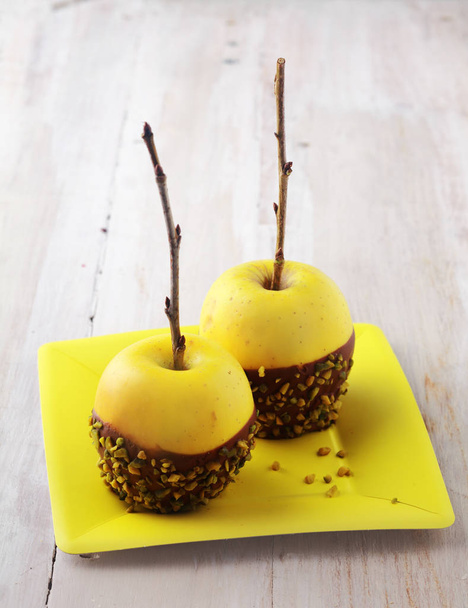 Healthy fresh organic golden yellow apples decorated with chocolate and chopped nuts served on a square yellow plate for a Halloween dessert, rustic white board background with copyspace - Photo, image