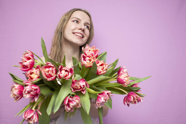 young beautiful girl with a bouquet of flowers on a colored pink background, a woman holds tulips and smiles - Foto, imagen