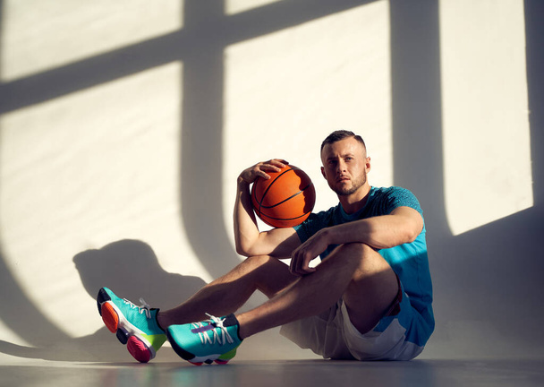 Joven atleta, jugador de baloncesto sosteniendo la pelota en el hombro y sentado cerca de la pared con sombras desde la ventana
 - Foto, imagen