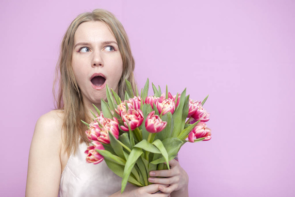 young surprised girl with a bouquet of flowers on a pink background, a cute woman holds tulips and rejoices - Photo, Image