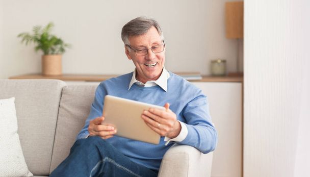 fröhlicher Senior mit digitalem Tablet sitzt auf Couch im Innenraum - Foto, Bild