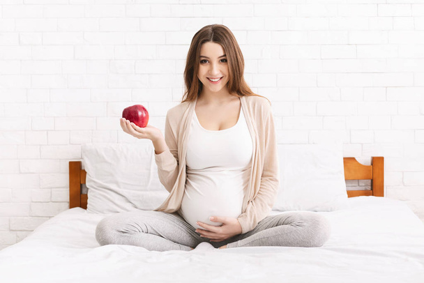 Young pregnant woman with red apple sitting in bed - Foto, Imagen