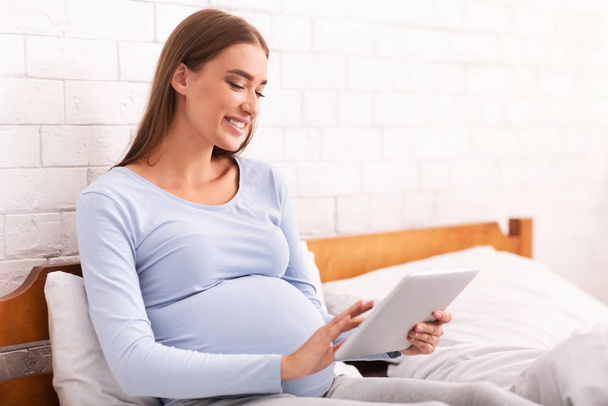 Щаслива вагітна дівчина використовує планшет сидячи в ліжку вдома
 - Фото, зображення