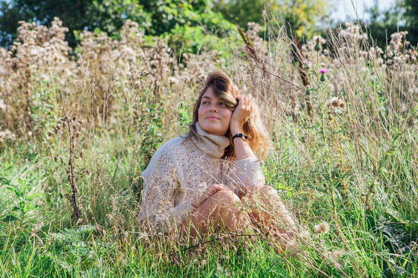 Happy young beautiful woman in sweater and underpants sitting with bare foot in a field of dry agrimony - Foto, afbeelding