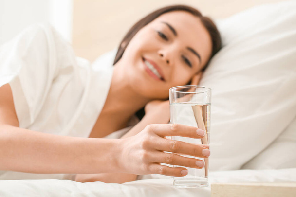 Ochtend van mooie jonge vrouw het nemen van glas water van nachtkastje - Foto, afbeelding