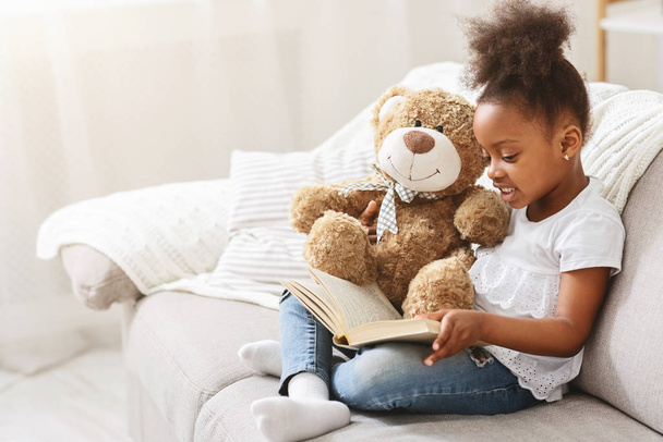 zwart klein meisje voorlezen sprookje naar haar teddybeer - Foto, afbeelding
