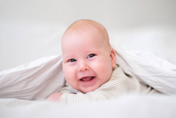 Adorable smiling baby boy under white blanket in bedroom. Newborn child relaxing in bed. Family morning at home. New born infant kids - Fotó, kép