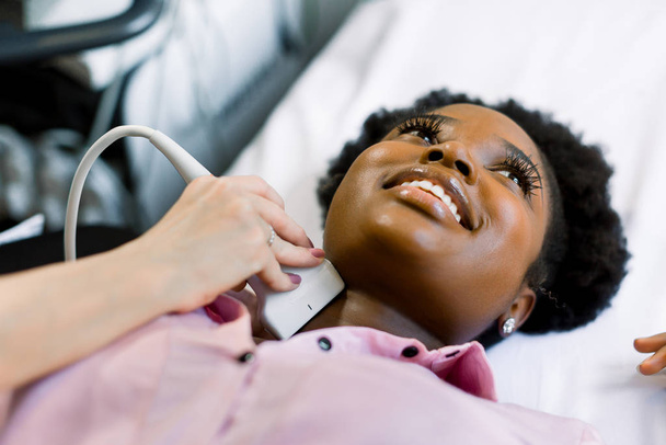 Close up shot of a young smiling African American woman getting her neck and thyroid gland examined by doctor using ultrasound scanner at the modern clinic - 写真・画像