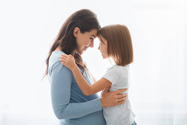 liebevolle schwangere Frau umarmt ihre kleine Tochter vor weißem Hintergrund - Foto, Bild