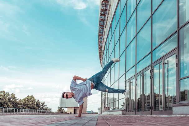 man stands on one arm, young guy dancer, in summer in city, dancing street dances, modern style of youth. Fashion and fitness sport. T-shirt glasses and jeans sneakers. Free space for copy text. - Photo, Image