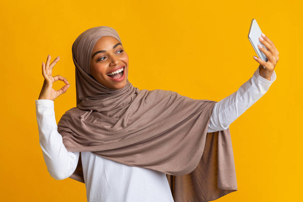 Cheerful black muslim girl in hijab taking selfie and gesturing ok - Fotoğraf, Görsel