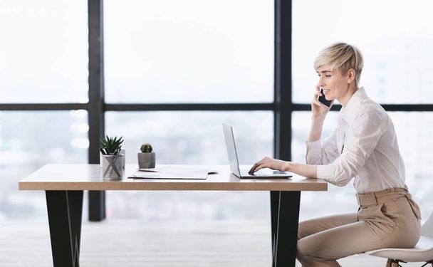 Femme d'affaires parlant au téléphone en utilisant un ordinateur portable dans le bureau, vue latérale
 - Photo, image
