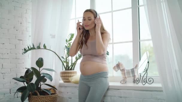 Smiling expectant mother listening music in headphones. Pregnant dancing at home - Video