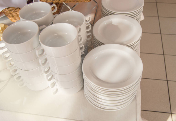 Lähikuva ryhmä valkoisia keraamisia teekuppeja ja lautasia, jotka seisovat pöydällä valkoisella pöytäliinalla, puhdista ruokia hotellin ravintolassa, joka on valmistettu vieraiden aamiaiseksi.
 - Valokuva, kuva